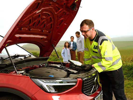 AA roadside assistance technician in a high-visibility yellow jacket inspecting the engine of a red car with the bonnet open on a rural roadside, while three people stand in the background watching.